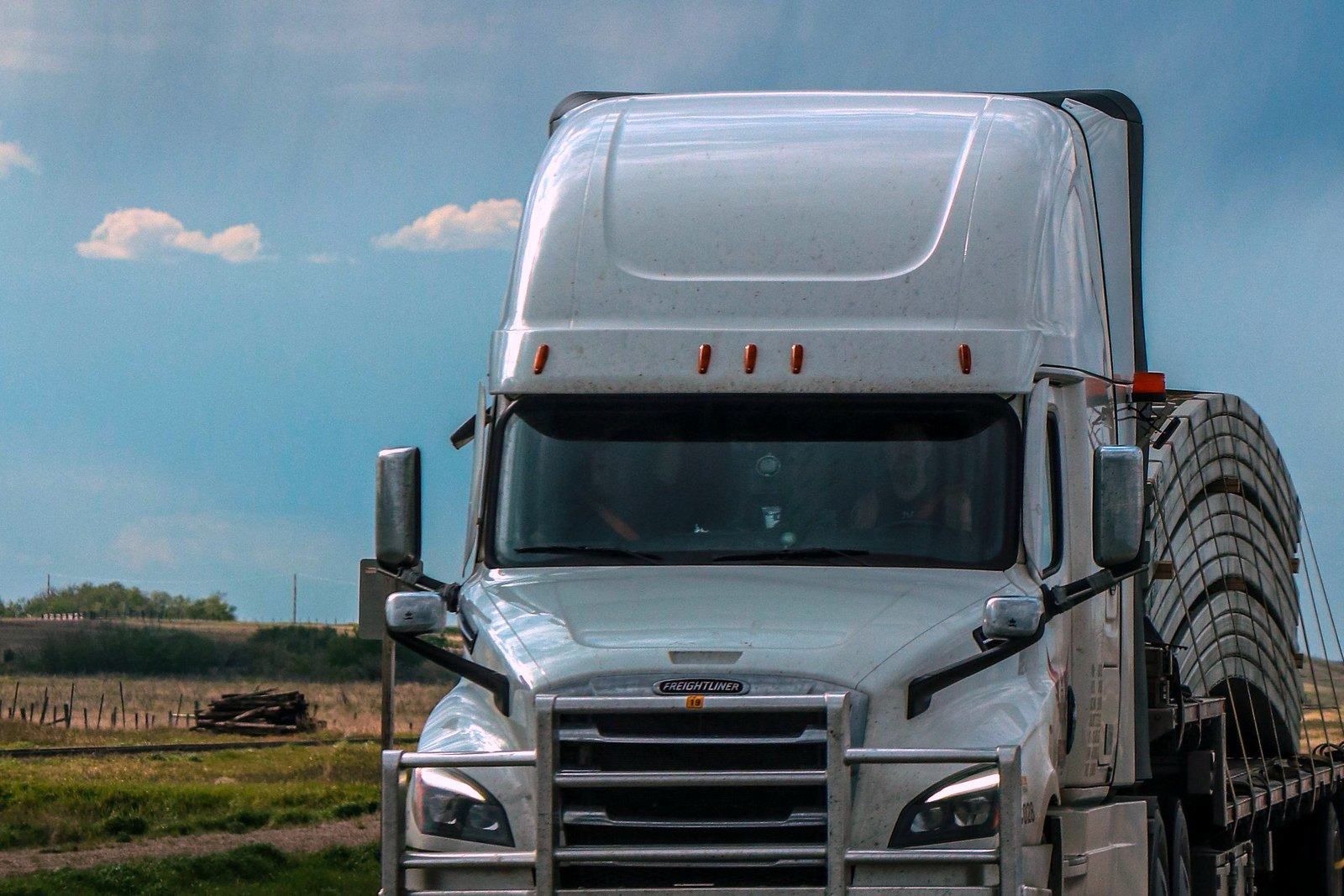 A white 18-wheeler hauling a tarped flatbed load along the edge of a flat, open Texas highway under a big sky