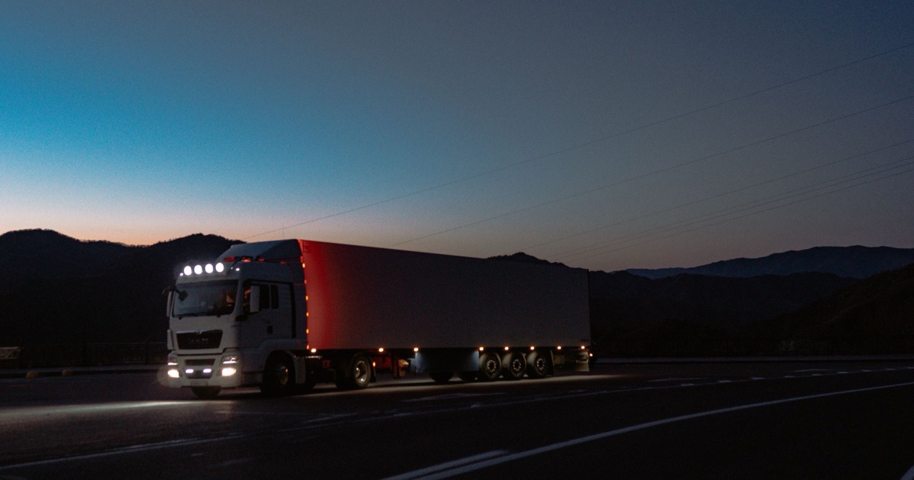 White semi truck with a red-lit trailer rolling down a mountain highway at twilight