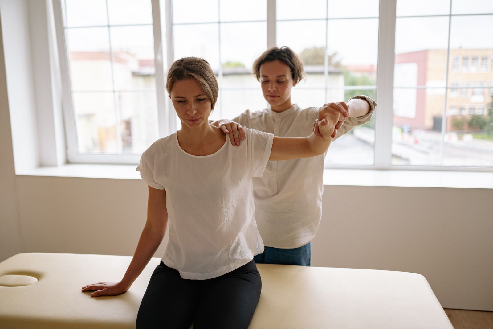 A physical therapist guiding a patient through arm and shoulder rehabilitation in a bright, window-lit therapy room